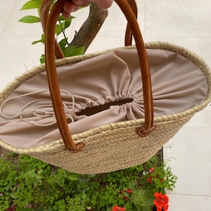 May include: A woven straw tote bag with brown leather handles. The bag has a beige fabric lining with a drawstring closure. The handles are attached with brown leather accents. The bag is oval-shaped and appears to be empty.