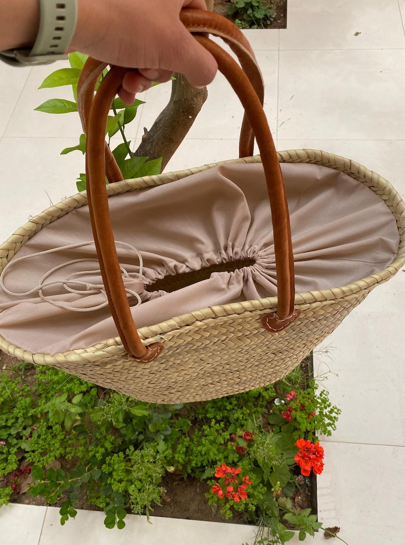 May include: A woven straw tote bag with brown leather handles. The bag has a beige fabric lining with a drawstring closure. The handles are attached with brown leather accents. The bag is oval-shaped and appears to be empty.