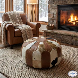 Peut inclure: Un pouf rond en cuir patchwork avec des sections marron, blanches et noires. Le pouf est dans un salon confortable avec un fauteuil en cuir, une couverture tricotée et une cheminée.