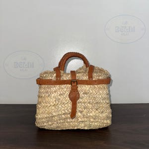 May include: Woven straw handbag with a brown leather handle and accents. The bag features a rectangular shape with a buckle closure and a woven texture. The bag is designed for carrying items.