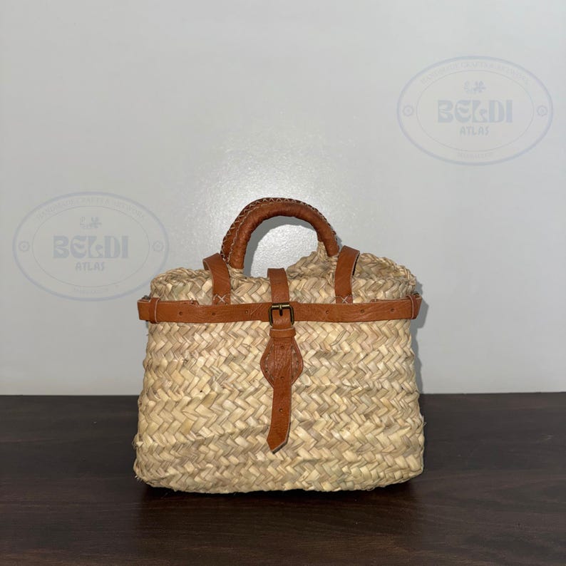 May include: Woven straw handbag with a brown leather handle and accents. The bag features a rectangular shape with a buckle closure and a woven texture. The bag is designed for carrying items.