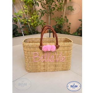 May include: Woven rectangular basket with brown leather handles and pink pom-poms. The words "Bella Vie" are embroidered in pink on the front. The basket is made of natural-colored woven material.
