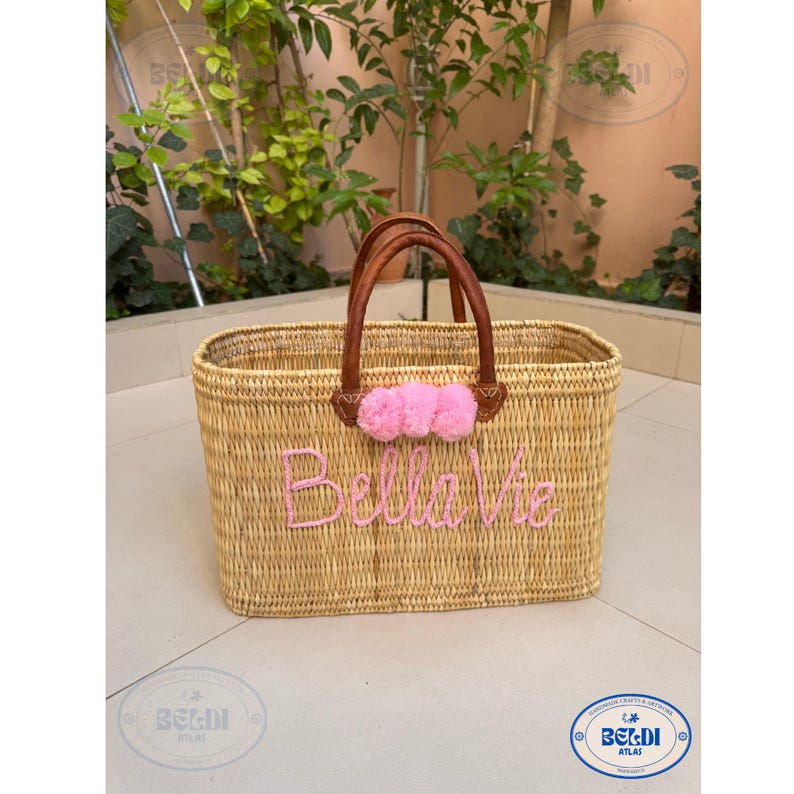 May include: Woven rectangular basket with brown leather handles and pink pom-poms. The words "Bella Vie" are embroidered in pink on the front. The basket is made of natural-colored woven material.