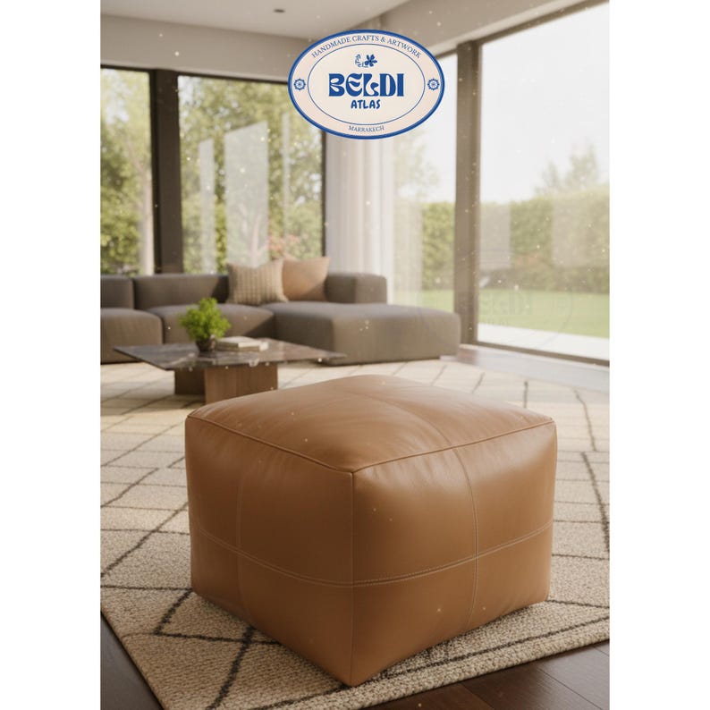 May include: A square, brown leather ottoman with stitched seams. The ottoman is placed on a patterned rug in a living room setting. The image also features a sofa, coffee table, and a window with a view of the outdoors. The text "BERDI ATLAS" is visible.