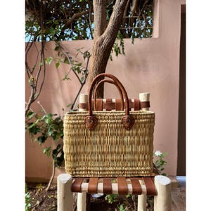 May include: A rectangular woven straw tote bag with brown leather handles and accents. The bag is sitting on a wooden chair with brown leather straps. The bag has a natural, light brown color.