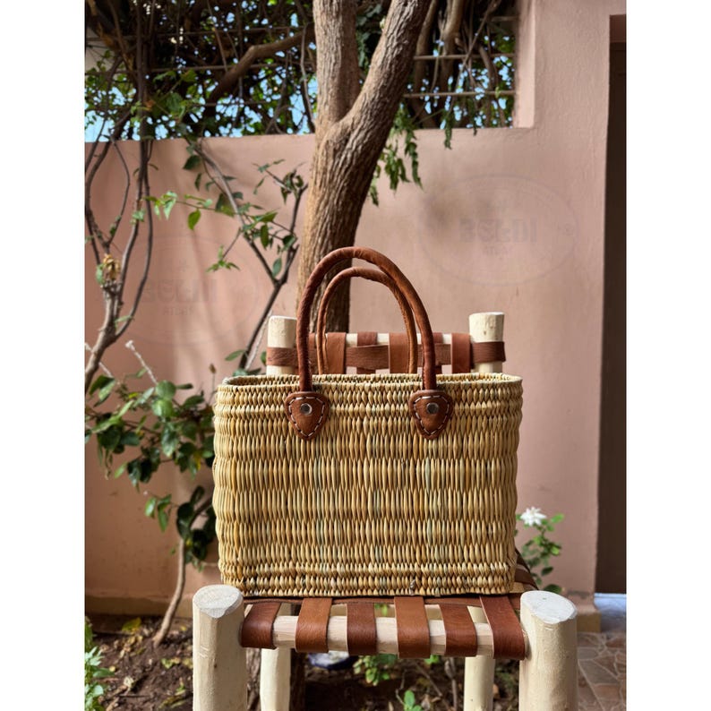 May include: A rectangular woven straw tote bag with brown leather handles and accents. The bag is sitting on a wooden chair with brown leather straps. The bag has a natural, light brown color.
