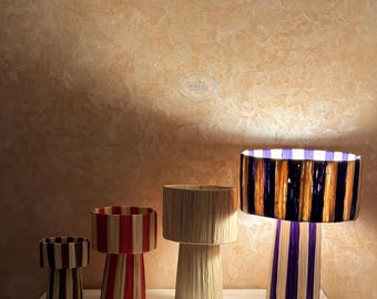 Könnte beinhalten: Eine Sammlung dekorativer Lampen mit gestreiften Schirmen in verschiedenen Größen. Die Lampen weisen eine Mischung aus Farben auf, darunter Rot, Weiß, Braun und Lila. Die Lampen sind auf einer weißen Oberfläche vor einem strukturierten braunen Hintergrund angeordnet.