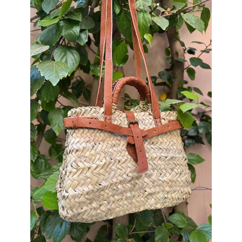May include: Woven straw tote bag with brown leather handles and a shoulder strap. The bag features a brown leather strap with a buckle closure. The bag is hanging in front of green foliage.