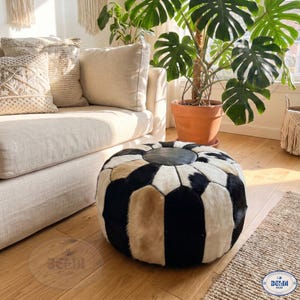 Könnte beinhalten: Ein runder Hocker aus Kuhfell in Schwarz, Weiß und Beige mit einer dunkelgrauen Lederoberfläche. Er steht auf einem Holzboden, in der Nähe eines beigen Sofas und einer großen Topfpflanze. Der Hocker hat einen Durchmesser von etwa 70 cm.