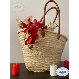 May include: A woven straw tote bag with brown leather handles and accents, filled with red and white flowers. The bag has a rounded shape and is sitting on a dark surface. Two red candles and a white candle are on the surface.