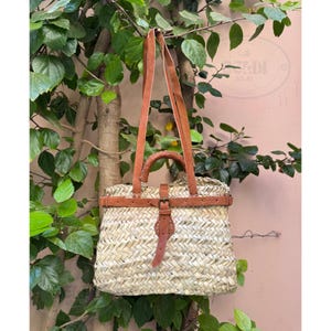May include: Woven straw tote bag with brown leather handles and accents. The bag features a rectangular shape with a buckle closure and a top handle. The bag is hanging from a tree branch.