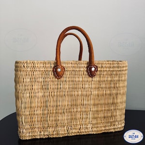 May include: A rectangular woven straw tote bag with brown leather handles. The handles are attached to the bag with brown leather patches and silver rivets. The bag is made of natural-colored straw.