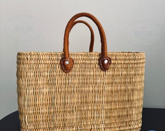 May include: A rectangular woven straw tote bag with brown leather handles. The handles are attached to the bag with brown leather patches and silver rivets. The bag is made of natural-colored straw.
