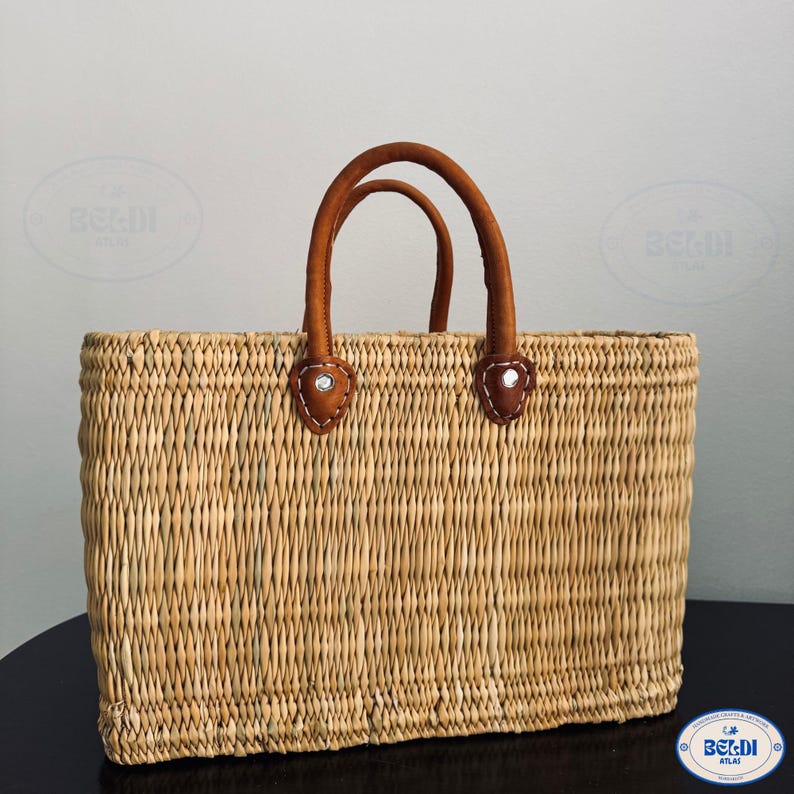 May include: A rectangular woven straw tote bag with brown leather handles. The handles are attached to the bag with brown leather patches and silver rivets. The bag is made of natural-colored straw.