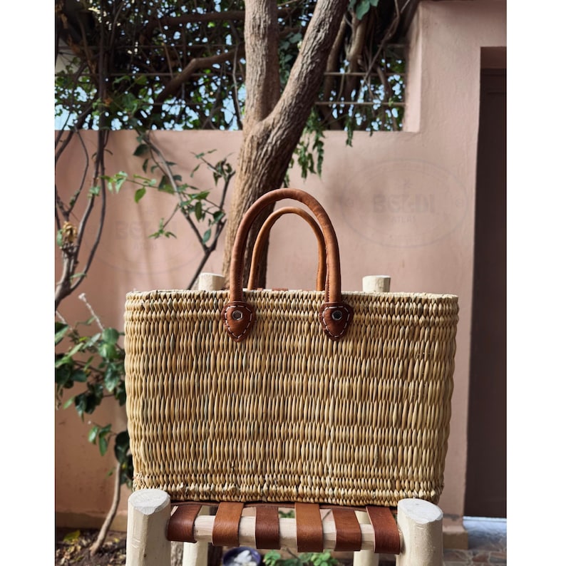 May include: A rectangular woven straw tote bag with brown leather handles and accents. The bag has a natural, light brown color and is sitting on a wooden bench with leather straps. The handles are arched and attached with leather patches.
