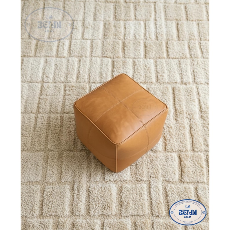 May include: A square, tan leather ottoman with stitched detailing. The ottoman is placed on a textured, off-white rug. The design is simple and modern, suitable for a living room or bedroom.