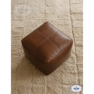 May include: A square, brown leather ottoman with stitched detailing. The ottoman is placed on a beige, textured rug. The design is simple and modern, suitable for various interior styles.