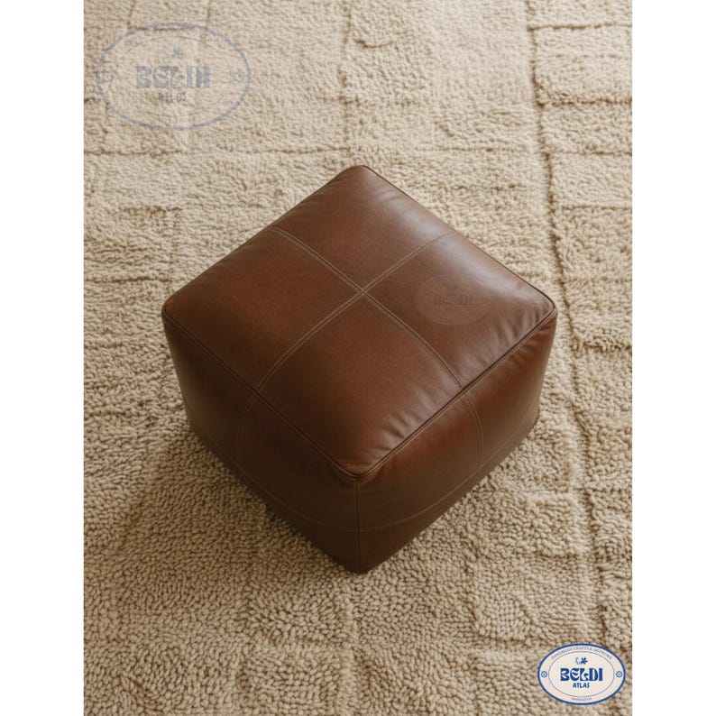May include: A square, brown leather ottoman with stitched detailing. The ottoman is placed on a beige, textured rug. The design is simple and modern, suitable for various interior styles.