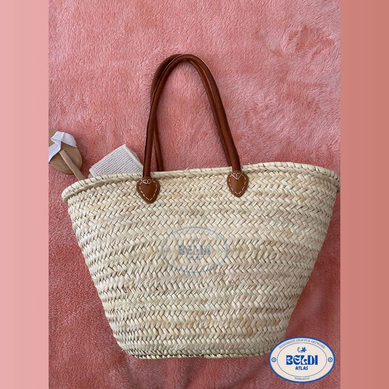 May include: Woven straw tote bag with brown leather handles and accents. The bag has a spacious interior and is ideal for carrying beach essentials. The bag has a logo on the front. The bag is sitting on a pink surface.