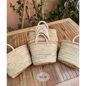 Op de afbeelding: Drie geweven strooien draagtassen met stevige handvatten worden gepresenteerd op een houten oppervlak. De tassen variëren in grootte en vorm, met een natuurlijke beige kleur. De handvatten zijn gemaakt van gedraaid touw. De tassen zijn van het merk Beldi Atlas.