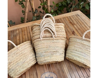 May include: Three woven straw tote bags with sturdy handles are displayed on a wooden surface. The bags vary in size and shape, with a natural beige color. The handles are made of twisted rope. The bags are from the brand Beldi Atlas.