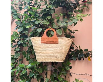 May include: A woven straw tote bag with a brown leather handle. The bag is rectangular with a rounded top and is set against a backdrop of green foliage and a pink wall. The handle is attached with visible stitching.