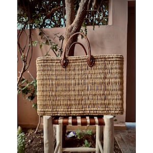 May include: A rectangular woven straw tote bag with brown leather handles and accents. The bag is sitting on a small wooden stool with a brown leather seat. The background includes a tree and a pink wall.