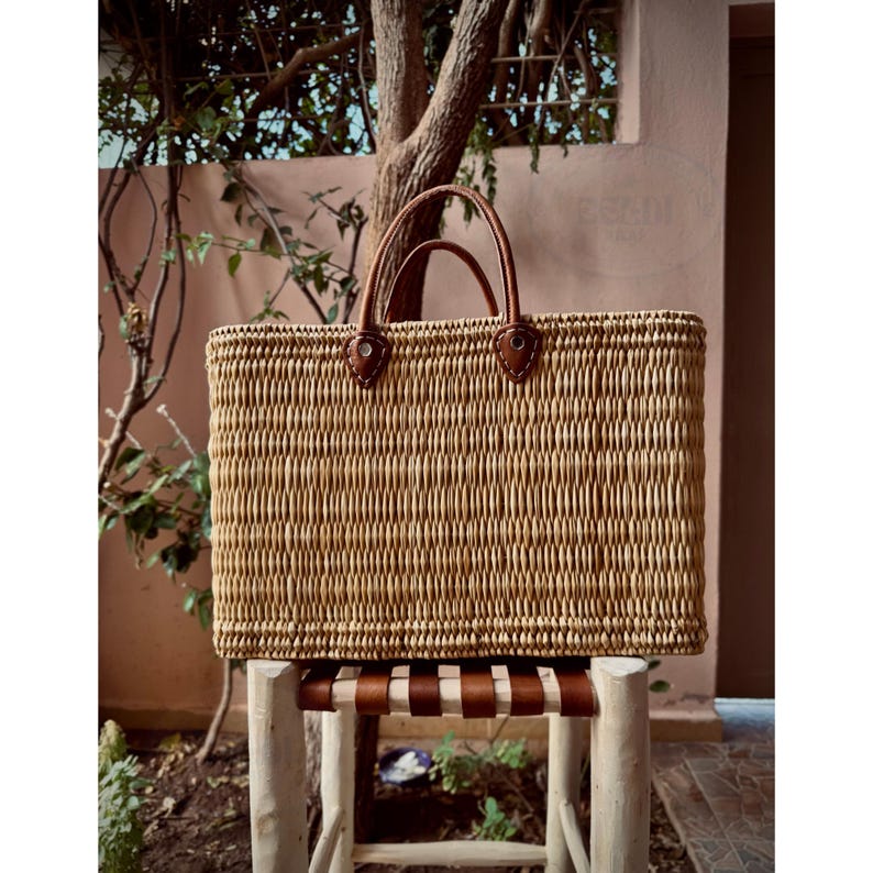 May include: A rectangular woven straw tote bag with brown leather handles and accents. The bag is sitting on a small wooden stool with a brown leather seat. The background includes a tree and a pink wall.