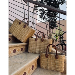 May include: Three rectangular woven baskets with brown leather handles are displayed on a staircase. The baskets are made of light brown woven material. The handles are attached with brown leather patches.