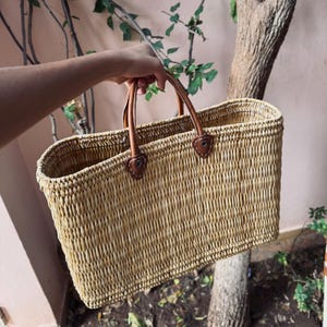 May include: A rectangular woven straw tote bag with brown leather handles and accents. The bag is held up by a person's hand. The bag is a natural beige color and appears to be empty.