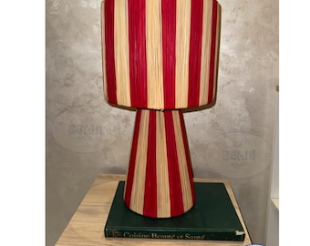 May include: A decorative lamp with a cylindrical shade and a conical base. The lamp features alternating vertical stripes of red and natural beige. The lamp is placed on a wooden surface, resting on a green book.