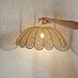 Könnte beinhalten: Eine dekorative Pendelleuchte mit offenem Gewebe und gewelltem Rand. Das Licht besteht aus einem Material in Naturtönen und wirft komplizierte Schattenmuster. Die Leuchte wird von einer Hand gehalten, die ihre Größe und ihr Design zeigt.