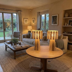 May include: Two wooden table lamps with striped shades sit on a round wooden table. The lamps are lit, casting a warm glow. The room features a neutral color palette, a sofa, and a coffee table.