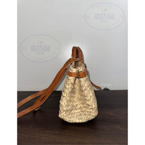 May include: A woven straw handbag with a brown leather handle and shoulder strap. The bag has a drawstring closure and a buckle detail. The bag is made of natural straw and brown leather.