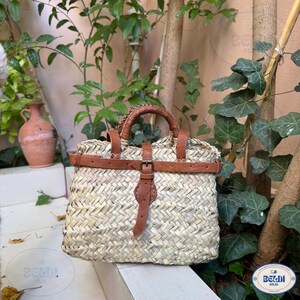 May include: Woven straw handbag with brown leather accents. The bag features a rectangular shape with a buckle closure and a leather handle. The bag is set against a backdrop of greenery and a terracotta vase.