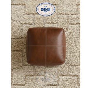 May include: A square, brown leather ottoman with white stitching details. The ottoman has a smooth surface and is placed on a textured, beige rug. The top of the ottoman has a stitched cross pattern. The image also includes a circular logo with the text "BELDI ATLAS".