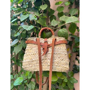 May include: Woven straw backpack with brown leather straps and handle. The bag features a rectangular shape with a flap closure and adjustable shoulder straps. The bag is set against a backdrop of green foliage.