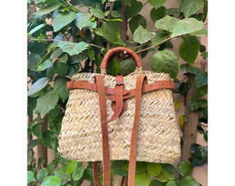 May include: Woven straw backpack with brown leather straps and handle. The bag features a rectangular shape with a flap closure and adjustable shoulder straps. The bag is set against a backdrop of green foliage.