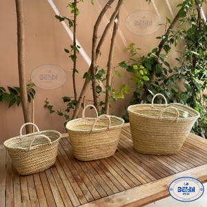 May include: Three oval-shaped woven straw baskets with handles in different sizes are displayed on a wooden table. The baskets are a natural light brown colour. The background features greenery and a light-coloured wall.