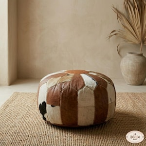May include: A round, low-profile ottoman constructed from panels of brown and white cowhide leather. The piece features a central circular patch with radiating rectangular segments, all joined by visible stitching. The brand name BELDI ATLAS is embossed.
