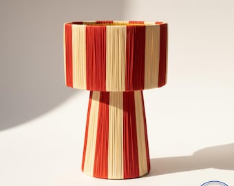 May include: A decorative lamp with a cylindrical shade and a conical base. The lamp features vertical stripes in alternating red and cream colors. The lamp is made of a woven material and stands on a white surface.