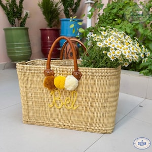 May include: A rectangular woven straw tote bag with brown leather handles and decorative pom-poms in brown, yellow, and white. The bag is filled with white and yellow flowers and has the word "Bel" embroidered in yellow.