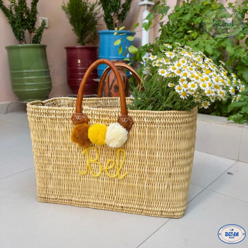 May include: A rectangular woven straw tote bag with brown leather handles and decorative pom-poms in brown, yellow, and white. The bag is filled with white and yellow flowers and has the word "Bel" embroidered in yellow.