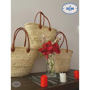 May include: Three woven straw tote bags with brown leather handles are displayed. A clear glass vase holds red and white flowers. Red and white candles are placed on a dark surface. The bags have a natural, light brown color.