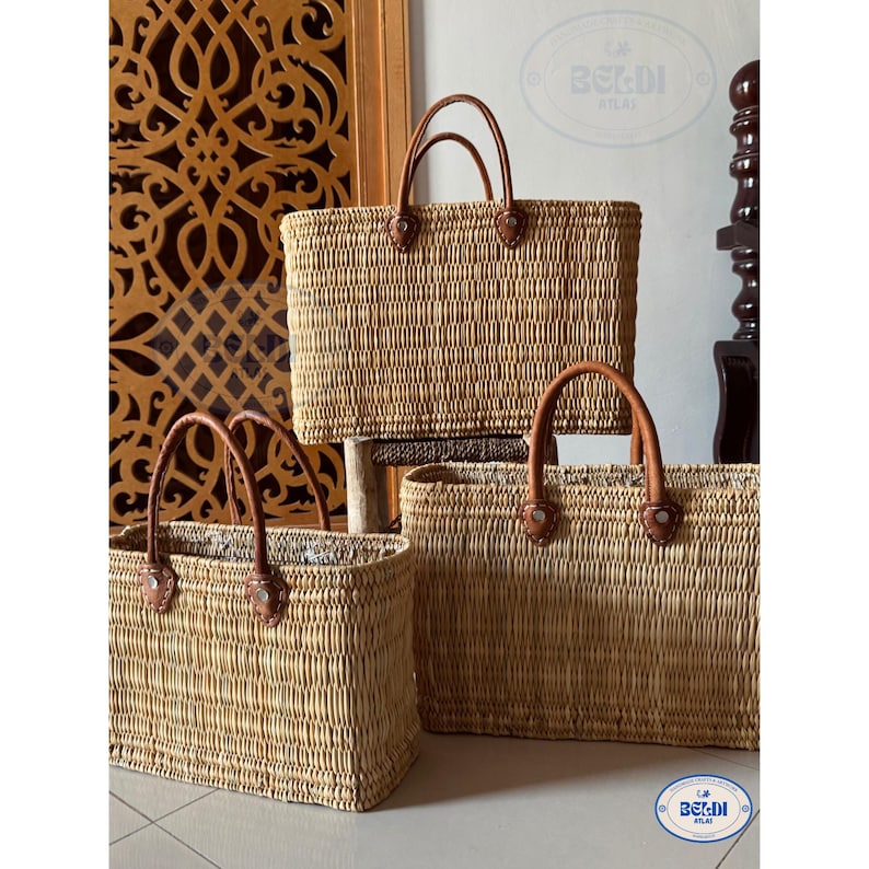May include: Three rectangular woven straw tote bags with brown leather handles. The bags vary in size, with the largest one in the background. The handles are attached with rivets. The bags are displayed on a light-colored surface.