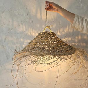 Könnte beinhalten: Gewebte Pendelleuchte mit konischem Schirm aus Naturstroh und Fransen. Das Licht wird von einer gedrehten Schnur gehalten und von einer Hand gehalten. Das Licht wirft einen warmen Schein.