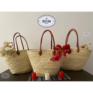 Puede incluir: Tres bolsos de mano de paja tejida con asas de cuero marrón. Las bolsas están llenas de flores y se colocan sobre una superficie oscura con velas. Las bolsas tienen el texto "BELDI ATLAS".