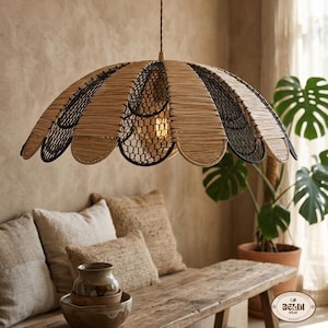Op de afbeelding: Een decoratieve hanglamp met een uniek, geschulpt ontwerp. De lamp heeft een geweven, natuurlijk gekleurde kap met zwarte accenten en een zwart gaasinterieur. Het licht hangt boven een houten bank met kussens en een plant in pot.