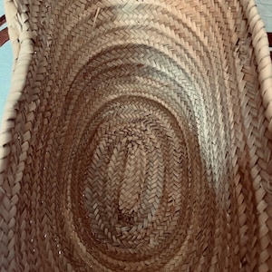 May include: A woven basket with a natural, light brown color. The basket has a rounded, oval shape and is made of tightly woven straw or similar material. The interior shows a spiral pattern, and the handles are not visible in this view.