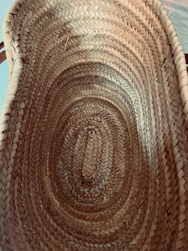 May include: A woven basket with a natural, light brown color. The basket has a rounded, oval shape and is made of tightly woven straw or similar material. The interior shows a spiral pattern, and the handles are not visible in this view.